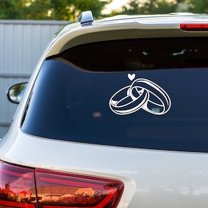 May include: A white car with a rear window sticker of two interlocking rings and a small heart above them.