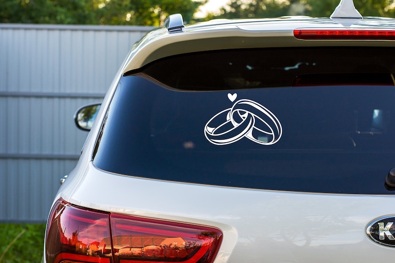 May include: A white car with a rear window sticker of two interlocking rings and a small heart above them.