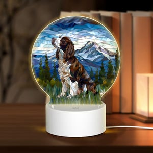 May include: A white night light with a stained glass design of a brown and white dog in a mountain landscape. The night light has a round base and a clear plastic dome.