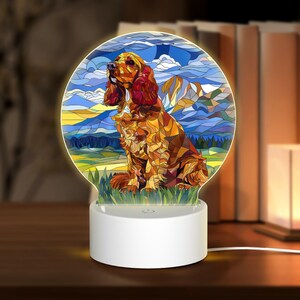 Könnte beinhalten: Ein weißes Nachtlicht mit einem runden, farbigem Glasdesign, das einen braunen und roten Cocker Spaniel Hund zeigt, der in einem Feld mit einer Bergkette im Hintergrund sitzt.