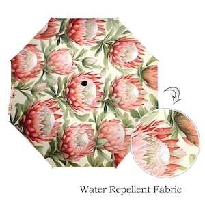 Puede incluir: Un paraguas rosa y verde con un estampado floral de proteas. El paraguas es repelente al agua y tiene un primer plano de la tela que muestra gotas de agua en la superficie.