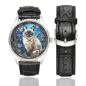 Op de afbeelding: Een zilveren polshorloge met een zwarte leren band heeft een glas-in-loodontwerp van een Siamese kat met blauwe ogen. De wijzerplaat van het horloge heeft zwarte cijfers en markeringen.