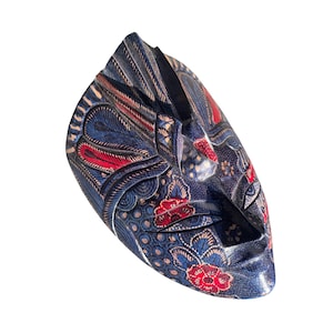 Op de afbeelding: Een blauw houten masker met rode en witte bloemenmotieven. Het masker heeft een gesneden gezicht met een gesloten mond en ogen.