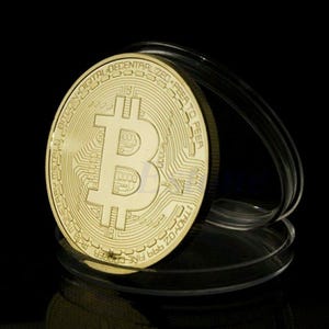 May include: A gold-colored Bitcoin coin with the text "Bitcoin Digital Decentralized Peer to Peer" around the edge. The coin is in a clear plastic display case.