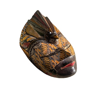 Op de afbeelding: Een handgesneden houten masker met een vogelachtig ontwerp. Het masker heeft ingewikkelde patronen in tinten zwart, bruin, geel en rood. De snavel van de vogel en de lippen zijn rood geschilderd. Het masker wordt waarschijnlijk gebruikt voor decoratieve doeleinden.