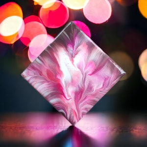 Peut inclure: Une peinture sur toile carrée avec des tourbillons abstraits roses, blancs et gris. La peinture est sur un fond sombre avec des lumières roses et blanches floues.