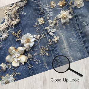May include: Close-up of a denim fabric adorned with delicate, handcrafted floral embellishments. The flowers are primarily white and cream, with intricate details and pearl accents. The text "Close-up Look" is visible.