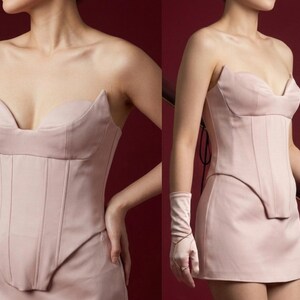 May include: A strapless, blush-colored corset dress with a structured bodice and a mini skirt. The dress features a sweetheart neckline and vertical seam detailing. The model is wearing matching gloves.