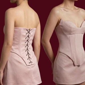 May include: A light pink strapless corset dress. The dress features a structured bodice with vertical seams and a mini skirt. The back of the dress has a lace-up closure with black lacing. The dress is shown from the front and back.