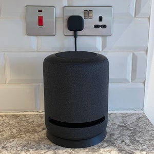 May include: A black cylindrical smart speaker with a fabric covering and a black base. The speaker is plugged into a wall socket.