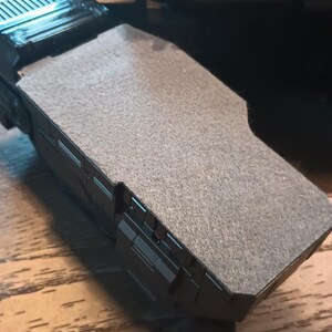 May include: A close-up of a black and grey toy spaceship with a grey felt-like surface on top.
