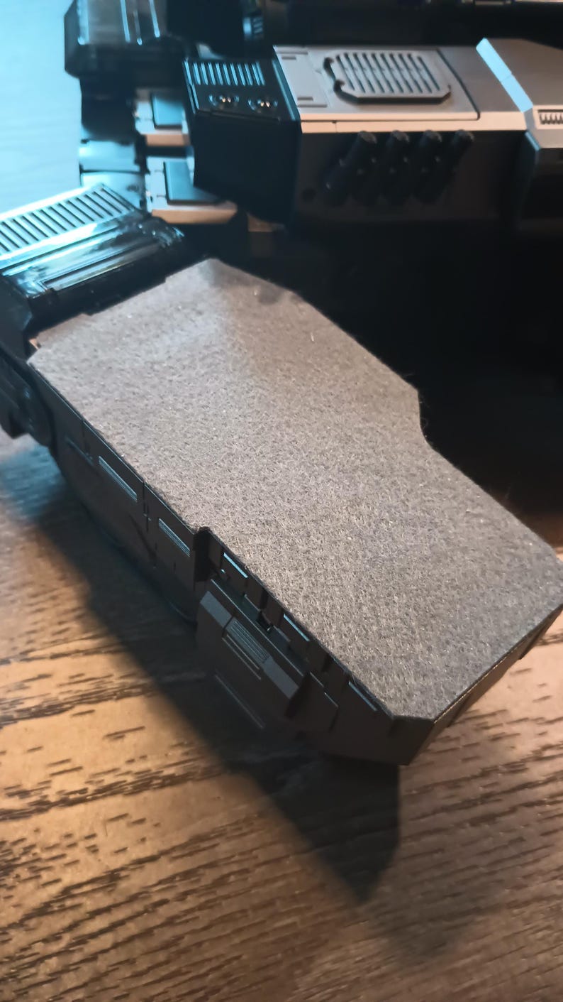 May include: A close-up of a black and grey toy spaceship with a grey felt-like surface on top.