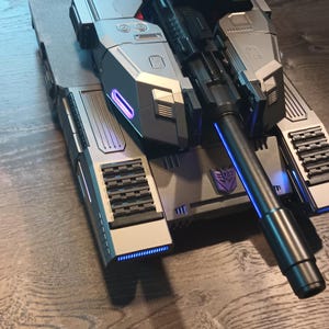 May include: A silver and black toy tank with a long black barrel and a purple Autobot symbol on the side. The tank has blue lights on the front and back.