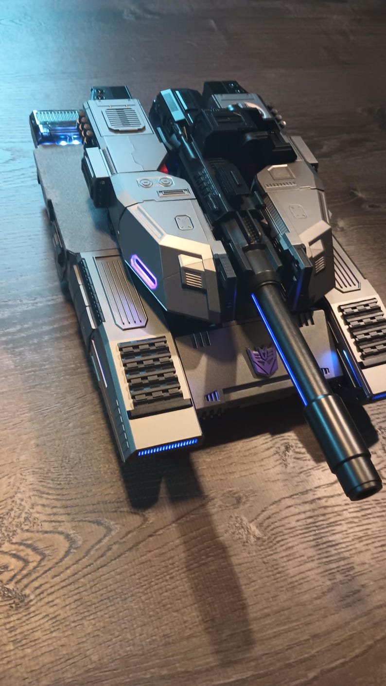 May include: A silver and black toy tank with a long black barrel and a purple Autobot symbol on the side. The tank has blue lights on the front and back.