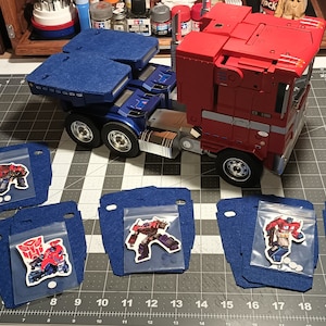 May include: A red, white and blue semi-truck toy with a blue trailer. The truck is surrounded by six blue felt squares, each with a clear plastic bag containing a different Transformers character sticker.