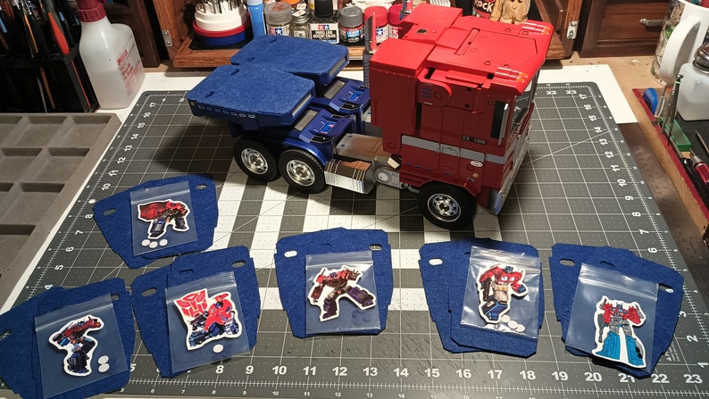 May include: A red, white and blue semi-truck toy with a blue trailer. The truck is surrounded by six blue felt squares, each with a clear plastic bag containing a different Transformers character sticker.