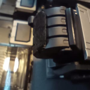 May include: Close-up of a black robotic hand with four fingers. The hand is made of metal and has a textured surface. The hand is attached to a larger robotic arm.