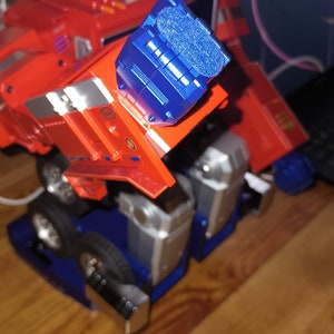 May include: A red and blue toy robot in the shape of a truck. The robot has a blue head and a blue arm. The robot is on a wooden surface.
