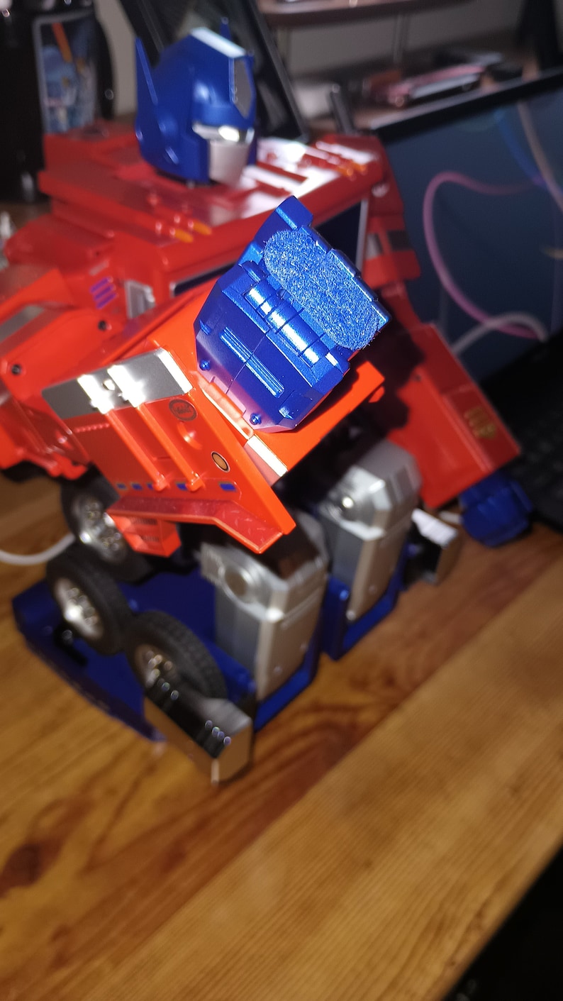 May include: A red and blue toy robot in the shape of a truck. The robot has a blue head and a blue arm. The robot is on a wooden surface.