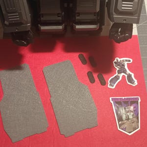 May include: Two grey felt floor mats with a textured surface, a sticker of a robot, and a sticker of a robot with the text "Transformers" on a red felt background.