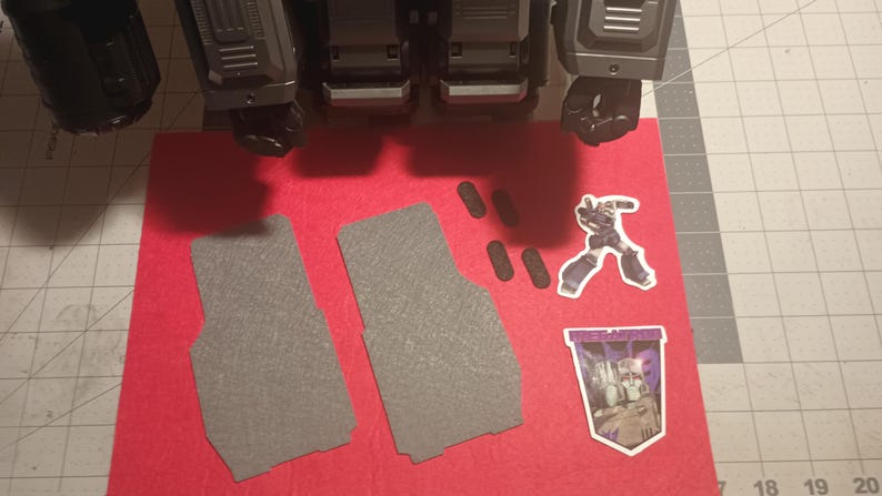 May include: Two grey felt floor mats with a textured surface, a sticker of a robot, and a sticker of a robot with the text "Transformers" on a red felt background.
