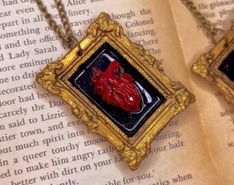 May include: A gold-framed necklace pendant with a red anatomical heart inside. The pendant is on a book with text.