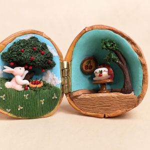 Puede incluir: Una cáscara de nuez con bisagras que contiene dos dioramas en miniatura. El lado izquierdo muestra un conejo rosa bajo un árbol verde con frutos rojos en un campo de hierba. El lado derecho muestra una mesa pequeña con un pastel, un árbol y un adorno de pared de cerezas.