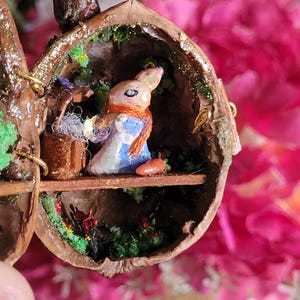 Pode incluir: Uma figurinha de coelho em miniatura vestindo um vestido azul e uma echarpe laranja senta-se dentro de uma casca de noz oca. A casca de noz é decorada com musgo verde, glitter e uma pequena prateleira de madeira. O coelho está segurando um pequeno objeto branco em suas mãos.