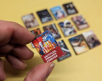 Puede incluir: Un libro en miniatura con una cubierta abstracta colorida, sostenido en una mano. El texto "UNTITLED, PERHAPS FOREVER" está en un fondo rojo. Varios otros libros en miniatura con diferentes cubiertas están en el fondo.