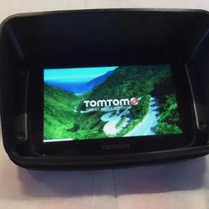 May include: A black TomTom GPS navigation device with a sunshade. The screen displays a scenic road with the text "TomTom Great Rides Edition".