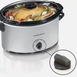 May include: A silver Hamilton Beach slow cooker with a black lid and handles. The slow cooker contains a cooked chicken with vegetables. The control knob is black and has the words "KEEP WARM" printed on the dial.