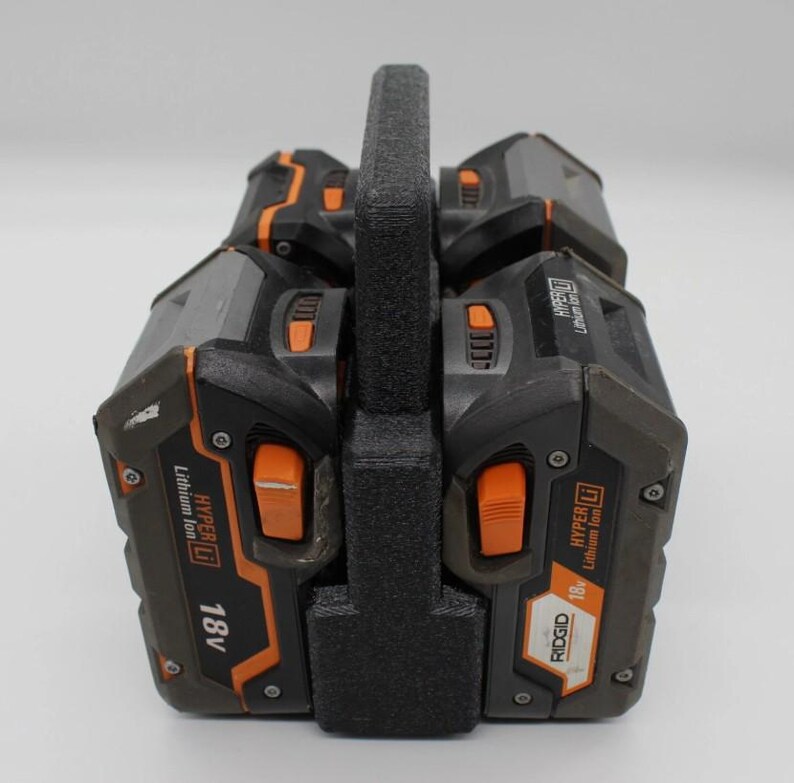 May include: A black and orange power tool battery holder with a handle. The holder contains four 18V Lithium Ion batteries. The batteries have orange buttons and the brand name "RIDGID" is visible.