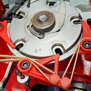 May include: Close-up of a mechanical device with a silver circular component at the center, surrounded by red levers and tan rubber bands. The device has a metallic appearance with a few visible numbers.