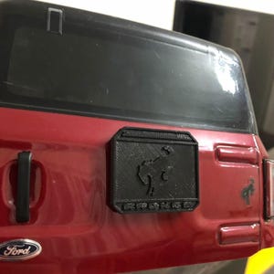 May include: A red Ford Bronco with a black plastic emblem on the back. The emblem has a running horse silhouette and the word "Bronco" written below it.
