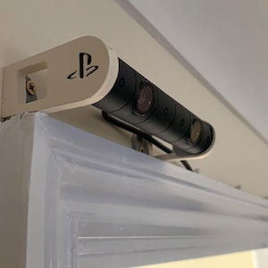 May include: A white and black PlayStation VR camera mounted on a white wall mount. The camera has three lenses and the PlayStation logo is visible on the mount.