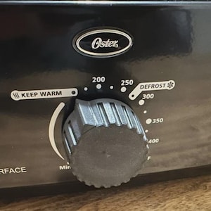 May include: Close-up of a black Oster appliance control panel. The dial has settings from Keep Warm to 400 degrees Fahrenheit, with a Defrost setting. The word "Oster" is above the dial.