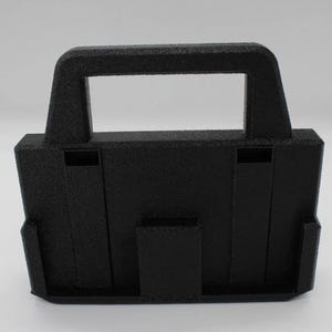 May include: A black, textured, 3D-printed carrying case with a handle. The case has a rectangular opening in the handle and a slot on the front. The case is designed to hold a small object.