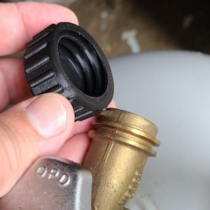 May include: A black plastic propane tank valve cover being held near a brass propane tank valve. The valve cover has a ridged exterior for grip. The valve is attached to a white propane tank.