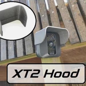 May include: A black plastic hood for a Blink XT2 security camera. The hood is designed to protect the camera from the elements and to improve its performance in direct sunlight. The hood is shown mounted on a wooden post.