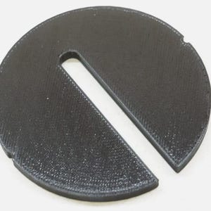 May include: A black plastic circle with a long, thin slot cut out of it. The circle has a textured surface.