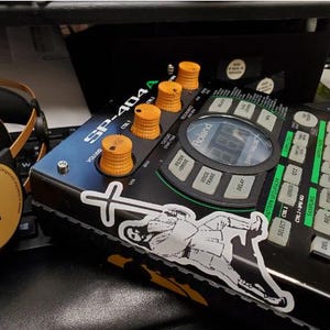 May include: A black Roland SP-404A sampler with orange knobs and a sticker of a person in a white robe with a cross on their back. The sampler has a variety of buttons and controls, including a display screen, a volume knob, and a filter knob.