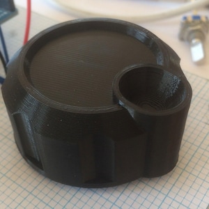 May include: A black plastic knob with a circular top and a cylindrical side. The knob has a textured surface and a small hole in the center.