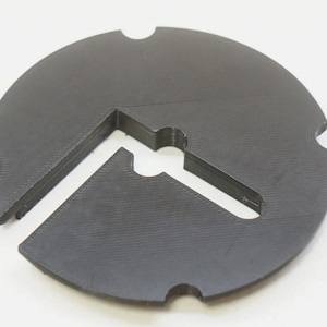 May include: A black plastic circular object with a cut-out shape in the center. The object has a textured surface and appears to be a mold or template.