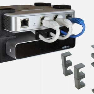 May include: A black 3D printed wall mount for a router and a streaming device. The mount has two shelves and includes cable management clips. The router and streaming device are not included.