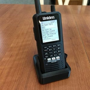 Puede incluir: Un radioescáner Uniden negro con una pantalla digital que muestra una lista de canales, incluyendo Lake County Public Safety, Lake County Law Enforcement Dispatch, County Sheriff/Police y Primary Dispatch. El radio está sentado en una base de carga negra.