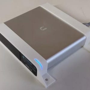 May include: A white wall mount for a silver device with a blue light. The device has a black front panel with the letter "U" on it.