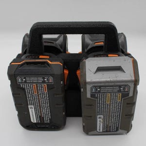 May include: A black and orange power tool battery charger with two battery slots. The charger has a black handle and the batteries have text that reads "PACK CHARGING RANGE" and "PACK RECHARGE GAMME".