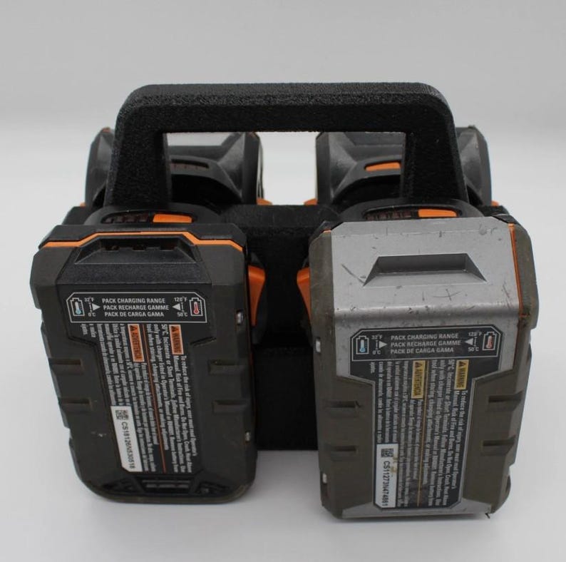 May include: A black and orange power tool battery charger with two battery slots. The charger has a black handle and the batteries have text that reads "PACK CHARGING RANGE" and "PACK RECHARGE GAMME".