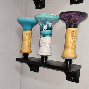 May include: Three ceramic hookah bowls with wooden bases mounted on a black wall shelf. The bowls are turquoise, teal, and purple with white and black designs. The bases are labeled "DOBAKO" and "OBMAKO".