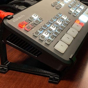 May include: A black ATEM Mini Pro video switcher with a gray control panel and white buttons. The switcher is mounted on a black stand.